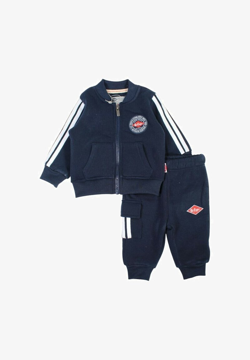 Survêtement de sport bleu marine pour enfants avec une veste à fermeture éclair ornée d'un écusson, un pantalon assorti avec une poche latérale et des bandes blanches sur les manches.