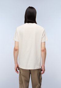 Person med langt mørkt hår iført en ensfarvet hvid kortærmet t-shirt og brune cargo-bukser, står vent imod en lys baggrund.