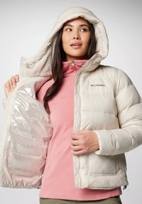 Lichte crèmekleurige pufferjack met een capuchon, voorzien van een glanzende voering, een gestikte ontwerpen en Columbia branding. Gedragen over een roze fleece top.