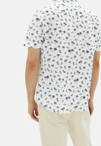 Camisa de manga corta hecha de tela ligera, con fondo blanco y patrones de palmeras y olas del mar en color azul marino.