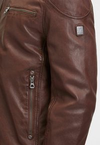 Braune Lederjacke mit einem Reißverschlusstasche, glatter Textur und einer kleinen Markenlogo-Plakette auf der Schulter.