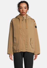Veste beige clair en matériau lisse avec capuche, cordon de serrage et deux poches latérales ; présente un accent texturé aux épaules et un écusson de logo.
