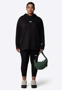 Sweat à capuche noir avec poche frontale, leggings noirs à motifs, sac à bandoulière vert et chaussures de randonnée beiges à lacets. Design simple et fonctionnel.