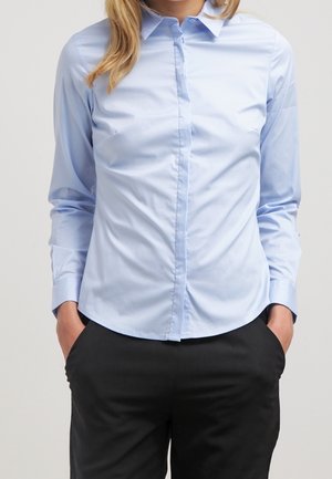 Lyseblå langærmet skjorte med knapper, båret uden at være stukket ned i sorte bukser, med hænderne i lommerne, foran en ensartet lys baggrund.