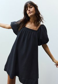Zwarte jurk met korte mouwen, een vierkante halslijn en gathered textuur. Heeft een losse pasvorm en een asymmetrische zoom.