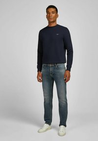 Homme portant une chemise à manches longues bleu marine, un jean bleu délavé et des baskets blanches se tenant devant un fond gris uni.