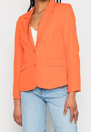 Vrouw draagt een fel oranje blazer over een witte top en lichtblauwe spijkerbroek, staand voor een egale achtergrond.