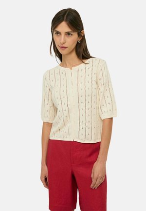 Mujer con un cárdigan de punto color crema con botones y mangas cortas y pantalones cortos rojos hasta la rodilla de talle alto, de pie con expresión neutral.