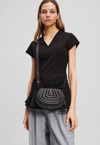 Borsa a tracolla nera con un motivo ondulato imbottito, forma rotonda e tracolla regolabile, abbinata a una maglietta nera e pantaloni grigi.