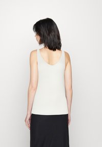 Rippenträgershirt in hellem Beige mit V-Ausschnitt auf der Rückseite. Weiche Textur mit taillierter Passform. Getragen mit einem schwarzen Rock.