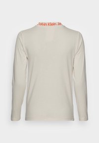Dlouhý béžový top s vysokým límcem a oranžovým nápisem Calvin Klein Jeans, zobrazující se zezadu na jednoduchém světlém pozadí.
