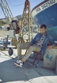 Informele outfit met een denim jack, een t-shirt met palmprint, kaki broek en zwart-witte sneakers, zittend op een houten stoel.