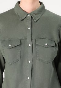 Chemise vert olive avec col, deux poches poitrine et boutons-pression argentés. Texture de tissu lisse avec coutures renforcées.