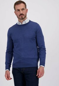 Donkerblauwe gebreide trui met een ronde hals, lange mouwen en geribbelde manchetten. Gedragen over een wit shirt met blauwe stippen.