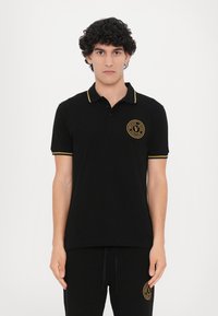 Polo negru cu un accent galben pe guler și mâneci. Prezintă un logo brodat rotund în zona stânga a pieptului.