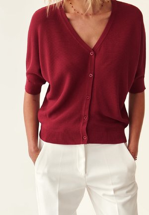 Frau trägt eine rote kurzärmelige, geknöpfte Strickjacke, kombiniert mit hoch taillierten weißen Hosen, Hände in den Taschen.