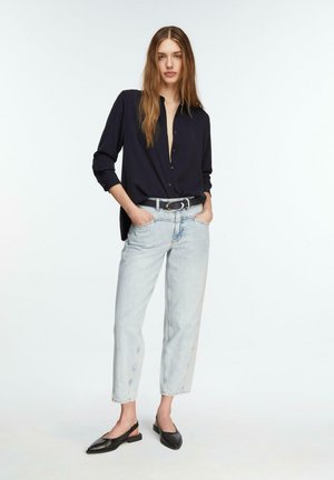 Frau steht mit den Händen in den Taschen, trägt ein schwarzes, geknöpftes Hemd, hellblaue Jeans, einen schwarzen Gürtel und schwarze Slingback-Ballerinas.