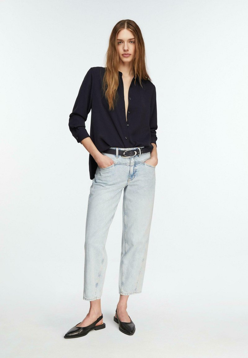 Vrouw staat met handen in haar zakken, draagt een zwart overhemd met knopen, lichtblauwe spijkerbroek, een zwarte riem en zwarte slingback platte schoenen.