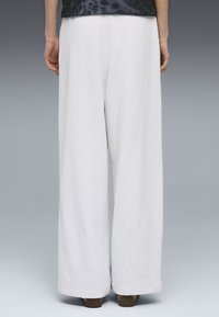 Άτομο που στέκεται, φοράει χαλαρά λευκά παντελόνια και σκούρα παπούτσια, εμφανίζεται από πίσω μπροστά σε απλό γκρι φόντο.