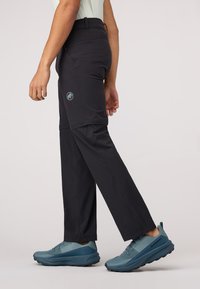 Pantalons noirs pour hommes, ajustés, conçus pour l'extérieur, en tissu léger, dotés d'un logo discret et de poches latérales. Associés à des chaussures de sport bleues.