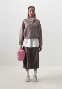 Sweatshirt castanho oversized com fecho, por cima de uma camisa branca, combinado com uma saia castanha plissada. A segurar uma mochila rosa da Nike. Sapatos e meias claras.