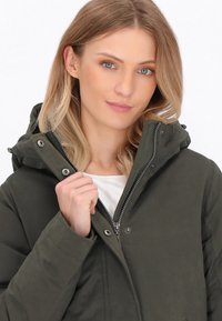 Žena s blond vlasy, oblečená v nezapnuté tmavě zelené bundě s kapucí přes bílé tričko, drží límec bundy jednou rukou.
