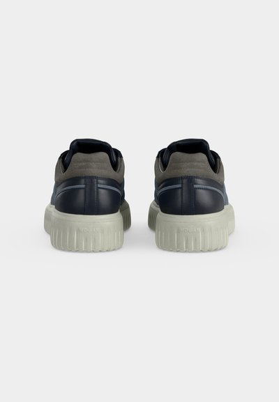 Paire de baskets bleu marine et grises avec des semelles blanches épaisses et texturées, vues de dos sur un fond uni gris clair.