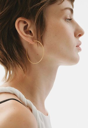 Pendiente de aro dorado con un diseño circular y delgado. Lleva puesto en la oreja izquierda de una modelo con el cabello corto, sobre un fondo blanco.