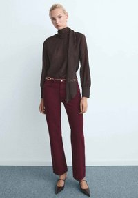 Femme portant un blouse à manches longues marron foncé avec un nœud au cou, un pantalon bordeaux, une ceinture en chaîne dorée et des escarpins marron à bout pointu.