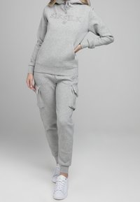 Sudadera gris claro y pantalones jogger a juego, confeccionados en una tela suave, con un logo en la sudadera y bolsillos tipo cargo en los pantalones. Combinado con zapatillas blancas.