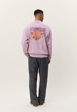 Vīrietis rozā džemperī ar basketbola grafiku un tekstu, valkājot pelēkas rūtainas bikses un brūnus apavus, stāvot ar muguru pret skatītāju.