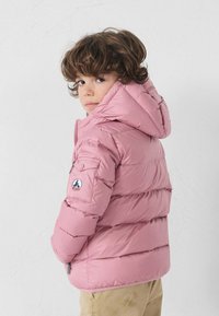 Chaqueta acolchada rosa con capucha, con costuras horizontales y un parche en la manga izquierda. El niño lleva pantalones color beige.