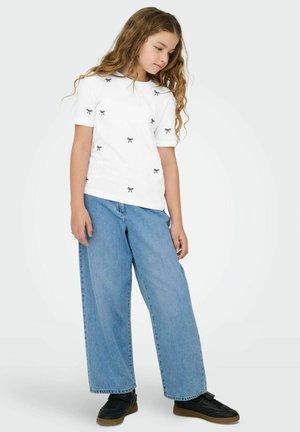 Ragazza con capelli lunghi e ricci che indossa una maglietta bianca con piccoli fiocchi neri, jeans larghi blu e sneakers nere, in piedi su uno sfondo bianco.