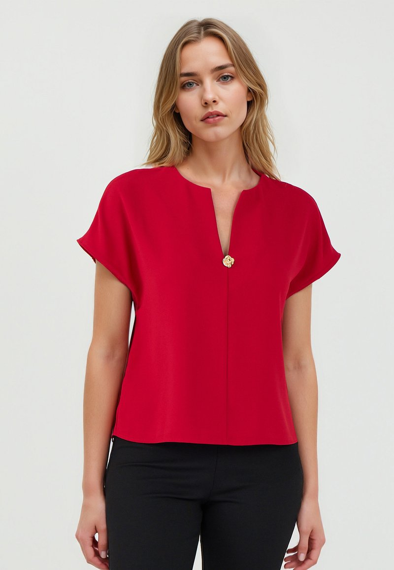 Blouse rouge à manches courtes avec un décolleté en V, dotée d'un bouton central en or. Tissu lisse avec une coupe décontractée et un ourlet droit.