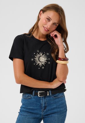Jeune femme aux longs cheveux bruns portant un t-shirt noir avec un motif soleil et lune, un jean bleu, et deux grands bracelets à son poignet.