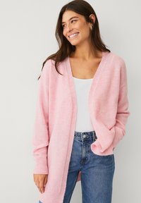 Cardigan tricoté rose clair avec manches longues, devant ouvert, poignets côtelés, et texture douce, porté sur un haut blanc et un jean bleu.