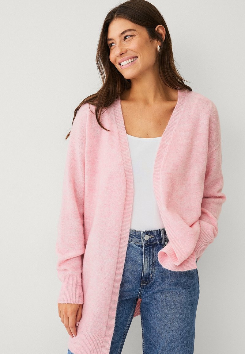 Cardigan tricoté rose clair avec manches longues, devant ouvert, poignets côtelés, et texture douce, porté sur un haut blanc et un jean bleu.