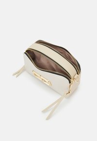 Petit sac bandoulière beige avec deux compartiments zippés ouverts, des dents de fermeture éclair dorées et un détail décoratif rectangulaire doré sur le devant.