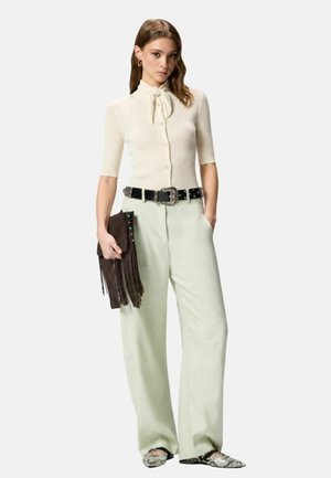 Donna che indossa un top crema a coste con fiocco al collo, pantaloni chiari a gamba larga, cintura nera, ballerine in pelle di serpente e tiene una pochette marrone con frange.
