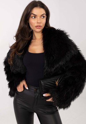 Schwarze Kunstpelzjacke mit langen Ärmeln, getragen über einem dunklen Oberteil, kombiniert mit hoch taillierten schwarzen Kunstlederhosen und einer gesteppten schwarzen Clutch.