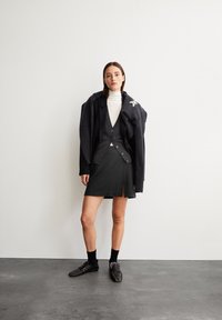 Frau trägt einen schwarzen Blazer, eine dunkelgraue Strickjacke, einen weißen Rollkragenpullover, einen schwarzen Rock mit Schlitz, schwarze genoppte Schuhe und einen dunklen Schal über den Schultern.