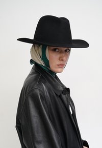 Schwarzer Filzhut mit breiter Krempe, getragen über einem grünen und beigen gemusterten Schal, kombiniert mit einer schwarzen Lederjacke. Glatte Textur und strukturiertes Design.