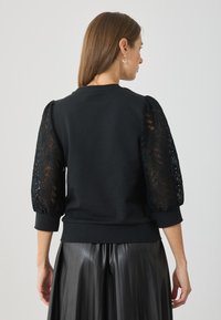 Sudadera negra con mangas abullonadas de encaje texturizado. Tejido del cuerpo suave y puños acanalados, con un diseño tipo pullover y cuello redondo.