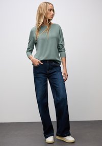 Hellgrüner Langarmpullover mit gerippten Bündchen, kombiniert mit weit geschnittenen dunkelblauen Jeans und weißen Sneakers vor einem grauen Hintergrund.