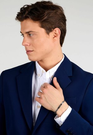 Blazer blu navy sopra una camicia bianca con bottoni. Bracciale argentato e nero sul polso. Texture morbide, design aderente, silhouette classica.