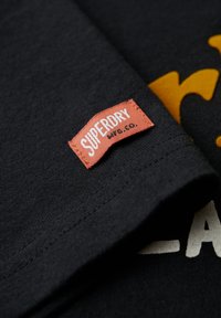 Czarny bawełniany T-shirt o fakturowanej tkaninie. Posiada wyraźną pomarańczową metkę oznaczoną "SUPERDRY" oraz odważne żółte grafiki.