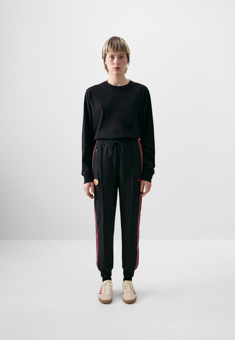 Chemise noire à manches longues et joggers noirs avec des rayures rouges sur les côtés, arborant un patch logo. Les chaussures sont crème avec des accents rouges.