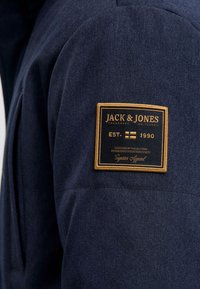 Temno modra jakna s teksturirano tkanino, izrazito zlato-črno znamčno značko na levem rokavu, z vtisnjenim napisom "JACK & JONES".