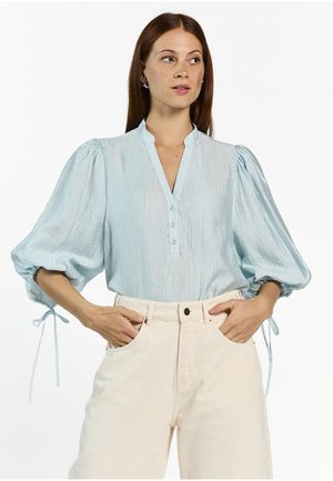 Vrouw met een lichtblauwe blouse met opgeblazen mouwen die bij de polsen zijn vastgebonden en crèmekleurige, high-waisted broek, handen in de zakken, zijwaarts kijkend.