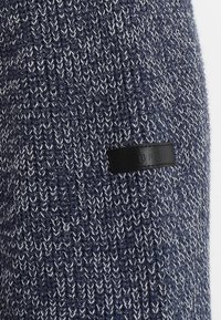Granatowy sweter z dzianiny o teksturowanym wzorze, z czarną skórzaną naszywką z logo w dolnym lewym rogu.